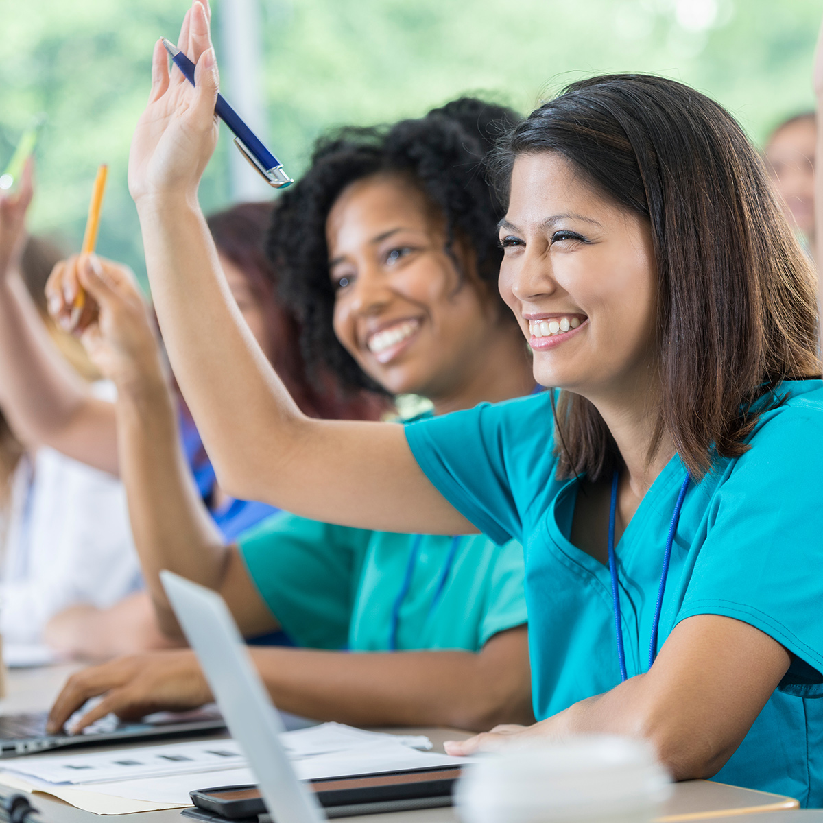 smiling women in scrubs in classroom setting, raising hands