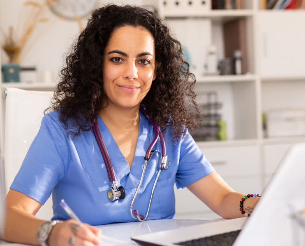 Female healthcare provider sitting at her desk in front of a laptop, holding a pen
