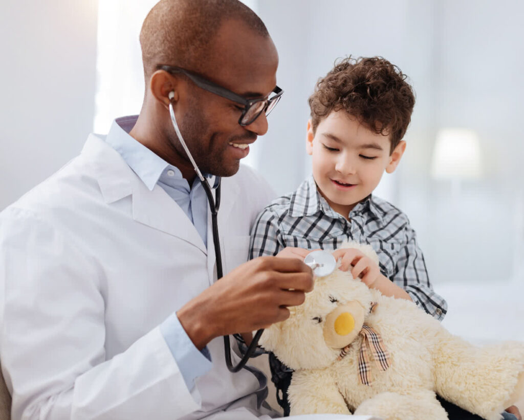 Male healthcare provider with young boy, using his stethoscope to examine a teddy bear